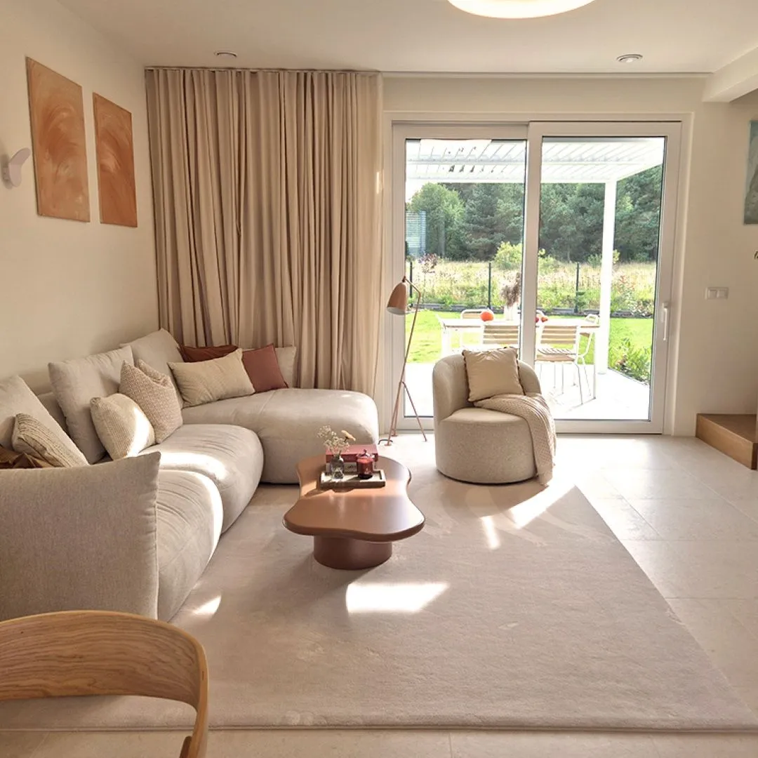 Salon w domu: kremowa narożna sofa, fotel bouclé, organiczny stolik, lniane zasłony i wyjście na taras z ogrodem